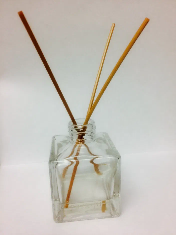Un diffuseur d’huiles essentielles composé de tiges de rotin et d’un flacon carré en verre, posé sur fond blanc.