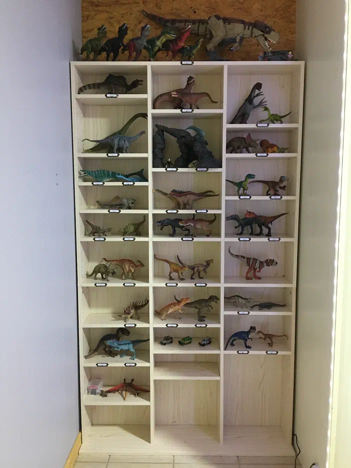 Une étagère en bois clair organisée en colonnes, remplie d'une grande variété de jouets dinosaures colorés et étiquetés.